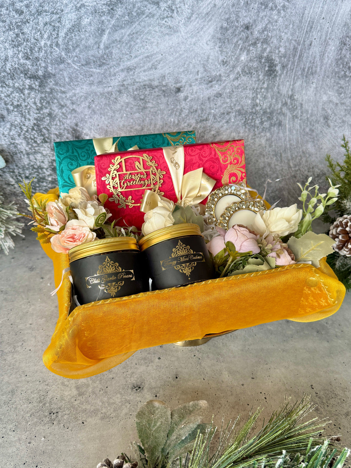 Gold holiday hamper with gift boxes, snack jars and flowers in a yellow fabric tray.
